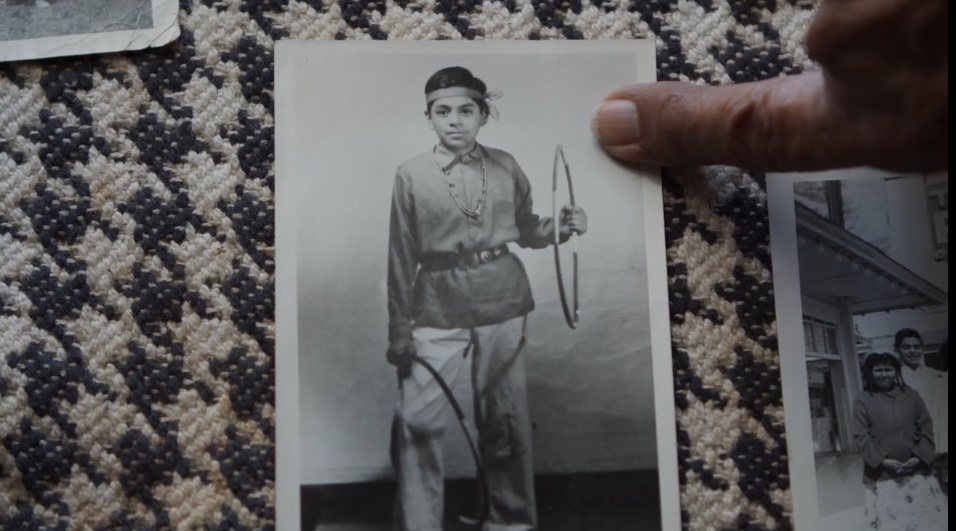 black and white photograph of a young Native boy holding a hoop in his left hand, and another hoop around his right leg. A finger is pointing at the photograph.